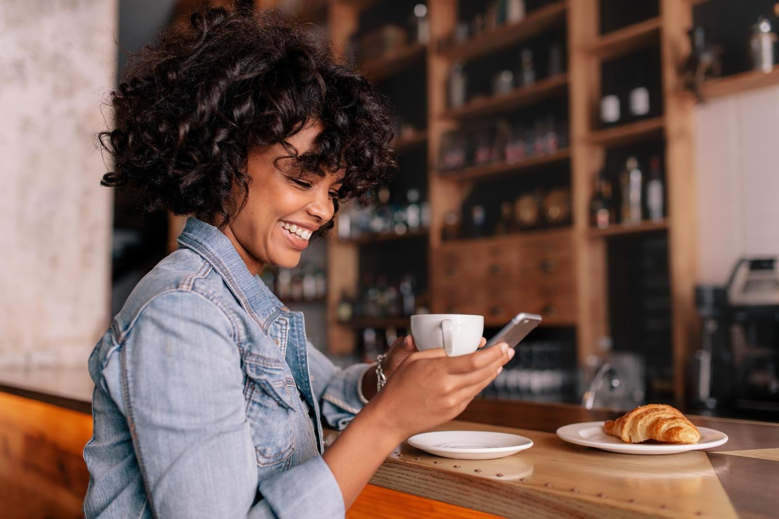 Smiling woman using smart phone in a modern cafe. Young african female sitting at a cafe counter having coffee and reading text message on mobile phone. Smiling woman using smart phone in a modern cafe